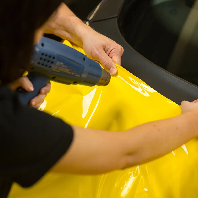 Installer using a heat gun to apply yellow vinyl wrap film to a vehicle